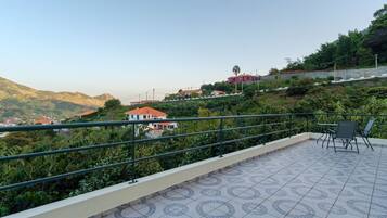 Eine Terrasse mit Fliesenboden, einem Metallgeländer und Blick auf einen Hang mit Gebäuden.