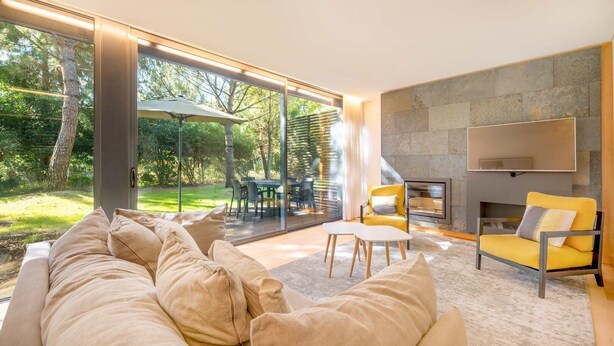 A modern living room with a sofa, yellow armchairs, a fireplace, and a glass door leading to an outdoor dining area. - GuestReady - Paradisiac Villa in Tróia (Praia de Troia)