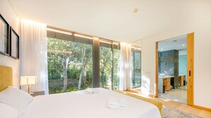A modern hotel room with a large bed, a bathroom with a sink and mirror, and a view of trees through large windows. - GuestReady - Paradisiac Villa in Tróia (Praia de Troia)