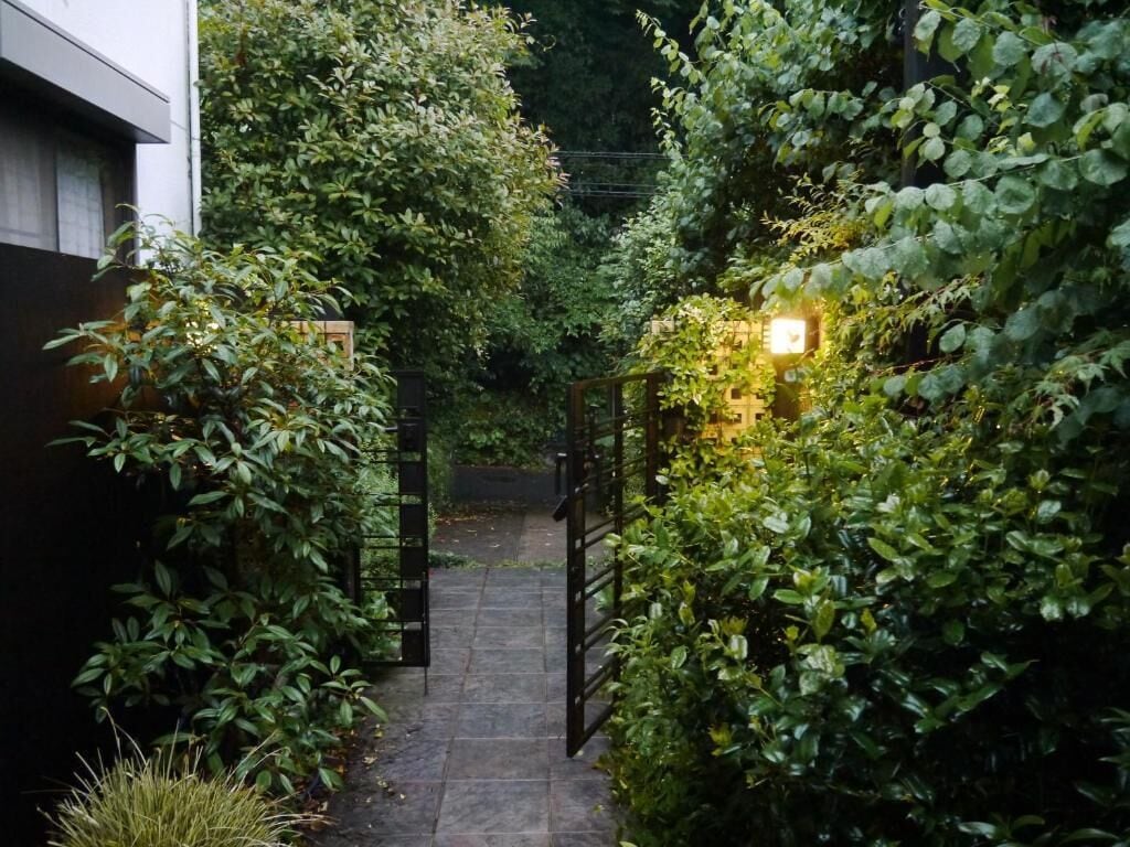 A narrow stone pathway flanked by dense foliage and a black gate, leading to a lit area.