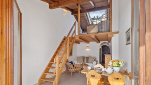 Una escalera de madera que conduce a un altillo con balcón, un comedor con sillas y mesa de madera, y un sofá al fondo.