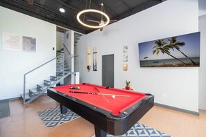 Ein Billardtisch in einem Raum mit Treppe, einem Strandbild und einem Deckenventilator.