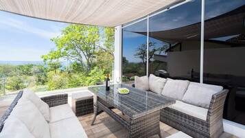 A modern outdoor seating area with a glass partition, wicker furniture, and a view of the landscape.
