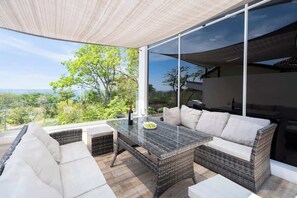 A modern outdoor seating area with a glass partition, wicker furniture, and a view of the landscape.