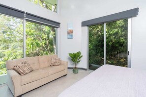 Ein modernes Schlafzimmer mit einem großen Bett, einem beigefarbenen Sofa, einem Blumentopf und einer Glastür zum Balkon.