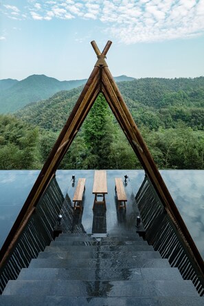 Estructura de madera con suelo reflectante que conduce a un área de estar con bancos y mesas, con vistas a una cadena montañosa boscosa.