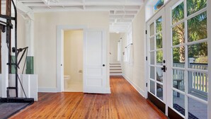 Un pasillo con suelo de madera, paredes blancas y una puerta de cristal que da a un espacio exterior con terraza de madera y palmeras.