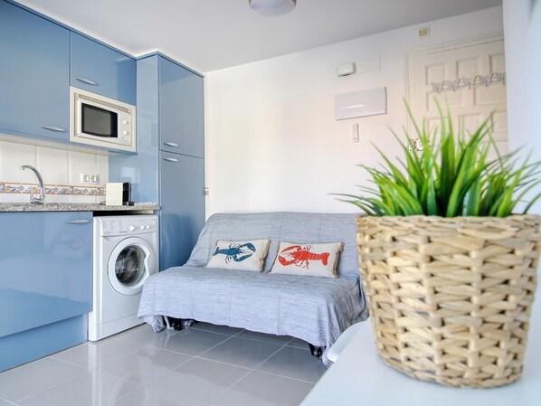 Cocina compacta con microondas, lavadora y un sofá gris con cojines decorados con langostas.