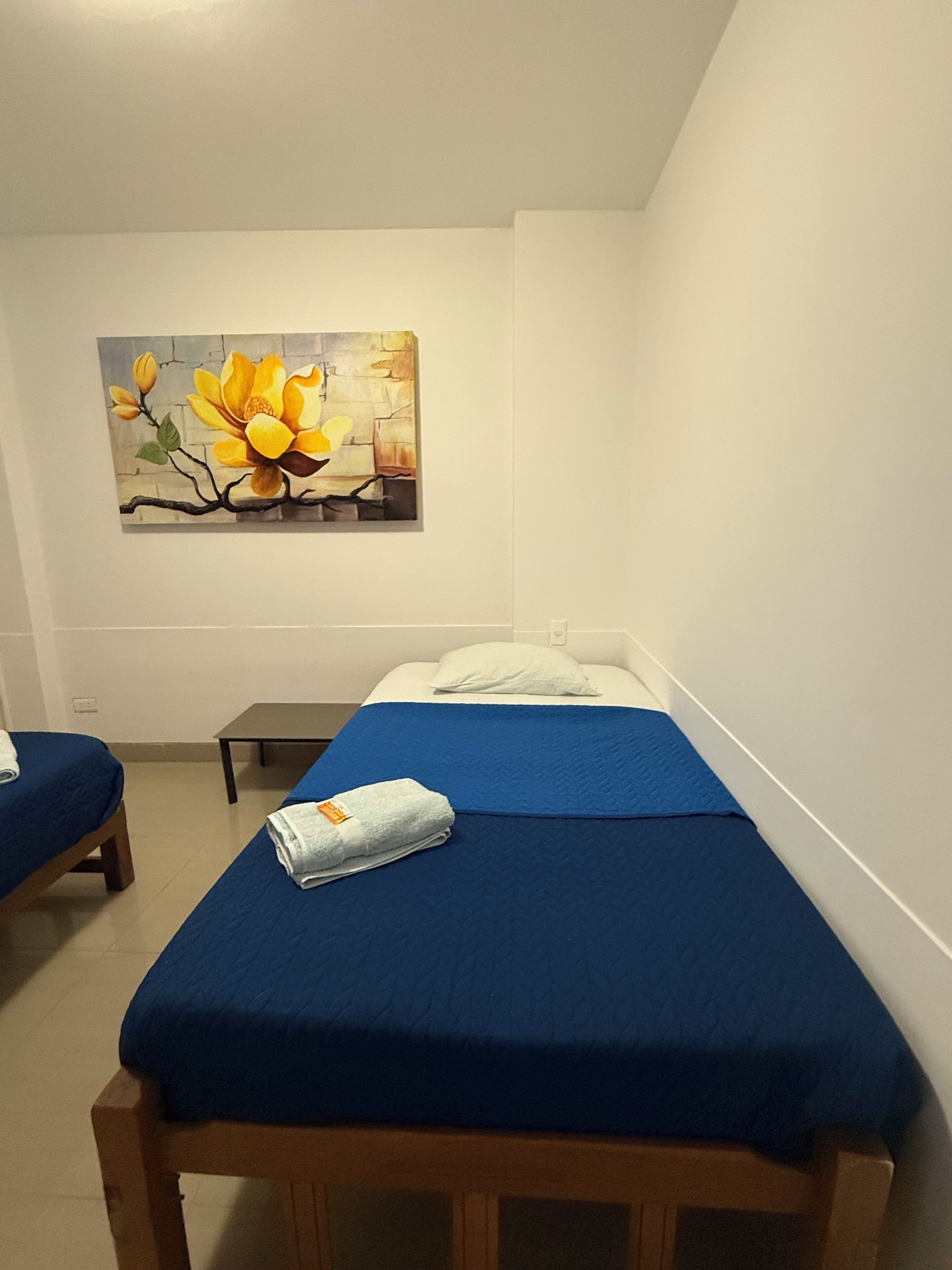 A single-bed room with a blue bedspread, a wooden bench, and a painting of yellow flowers on the wall.