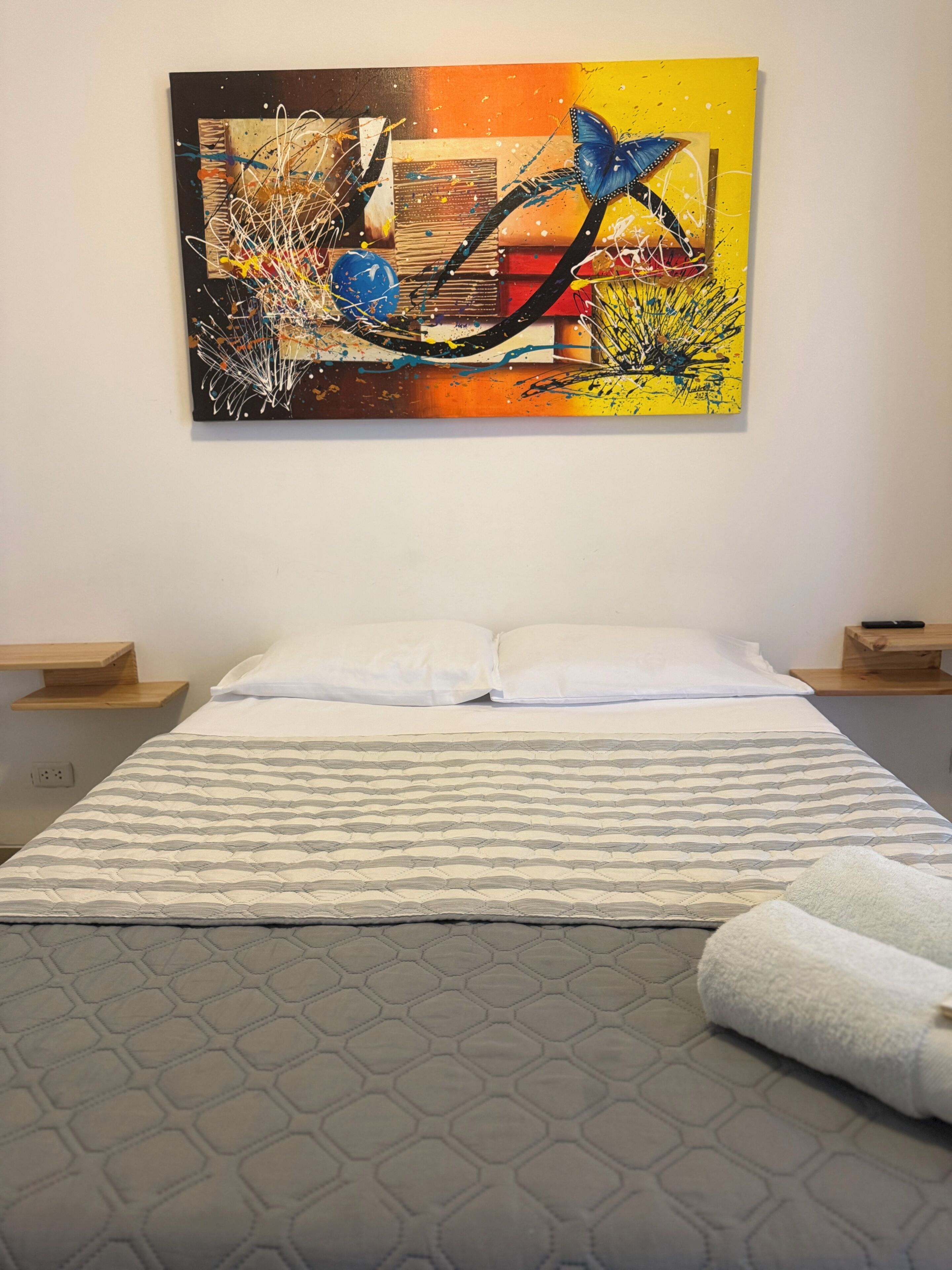 A bedroom with a bed, white pillows, a patterned bedspread, a wall-mounted abstract painting, and bedside tables.