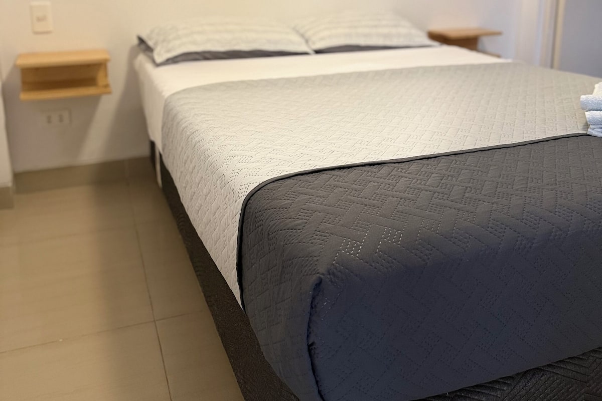 A neatly made bed with a white and dark grey quilt, a small bedside table, a wall-mounted painting, and a door to the right.