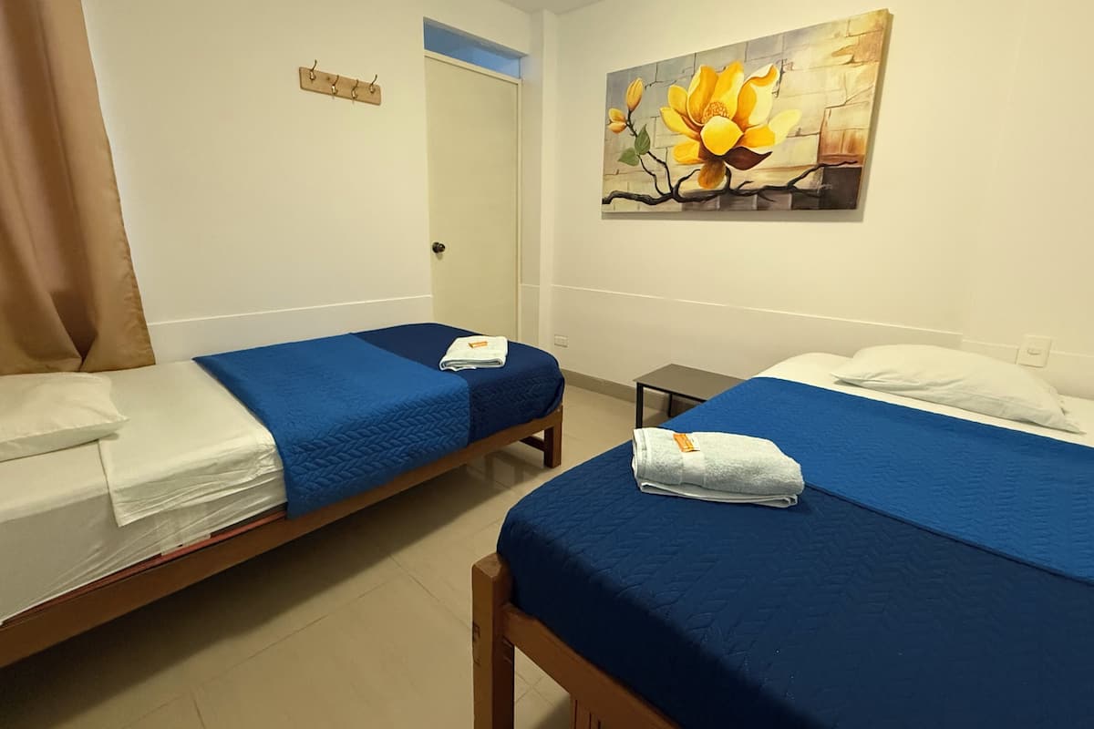 A hotel room with two single beds, each with a blue bedspread and white pillows. There is a painting of a flower on the wall, a small table, and a door with a towel hanging on the handle.