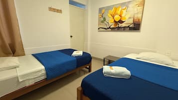 Habitación de hotel con dos camas individuales, cada una con una colcha azul y almohadas blancas. En la pared, un cuadro de una flor, una mesita y una puerta con una toalla colgando del pomo.