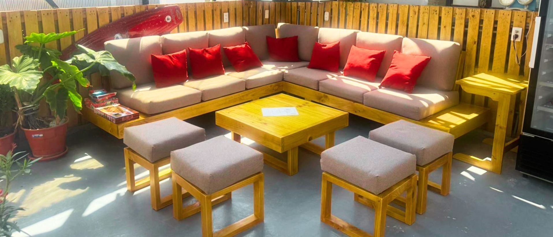 Quarto de hostel acolhedor com um amplo sofá em formato de L, duas mesinhas de madeira e uma mesa de centro. Há plantas em vasos e vista para o mar através de grandes janelas.
