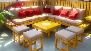 Quarto de hostel acolhedor com um amplo sofá em formato de L, duas mesinhas de madeira e uma mesa de centro. Há plantas em vasos e vista para o mar através de grandes janelas.