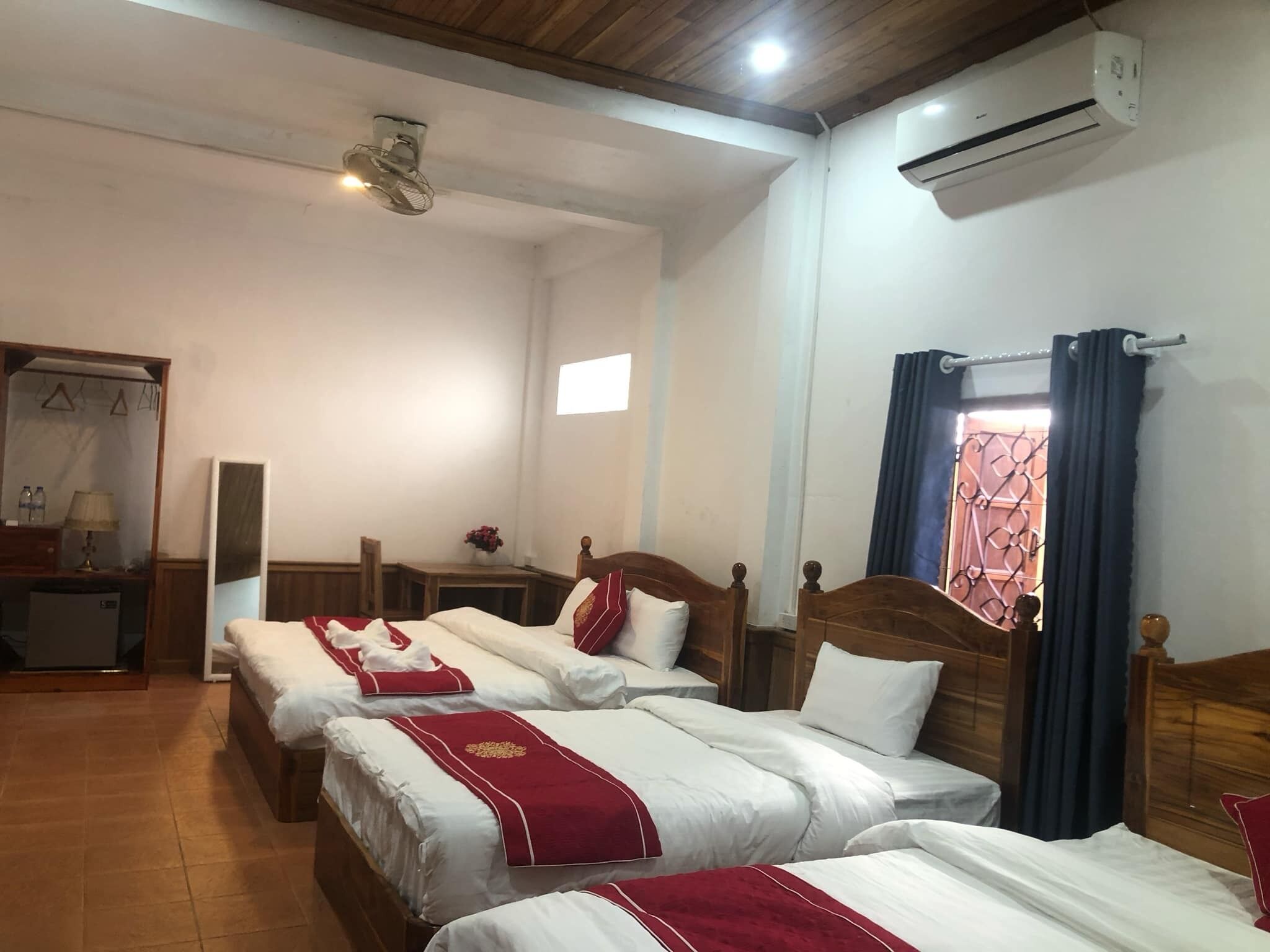 Ein Hotelzimmer mit drei Betten, Holzboden, Klimaanlage und einem mit Vorhängen versehenen Fenster.