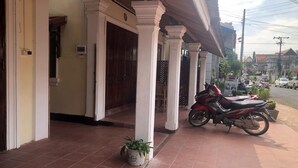 Ein Gebäudeeingang mit einer gefliesten Veranda, einem roten Motorrad davor und einem Blumentopf auf dem Boden.