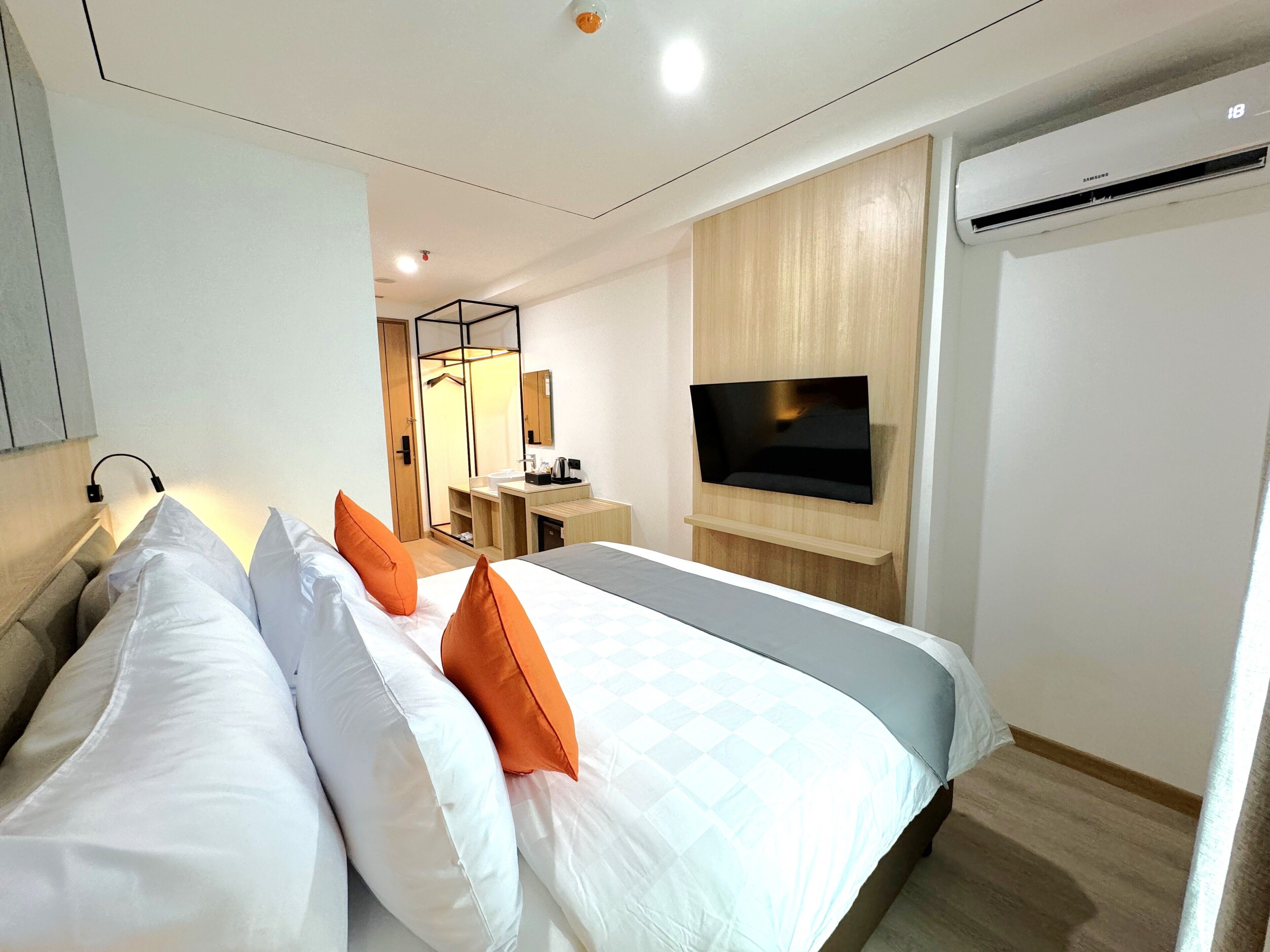 A modern hotel room with a large bed, a flat-screen TV mounted on the wall, a wooden wardrobe, and an air conditioning unit.