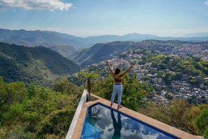 Eine Person steht auf einer Poolterrasse mit Blick auf eine Berglandschaft und eine Stadt darunter.
