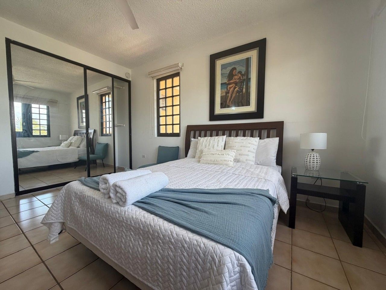 A hotel room with a bed, a bedside table with a lamp, a large mirror, and a painting on the wall.