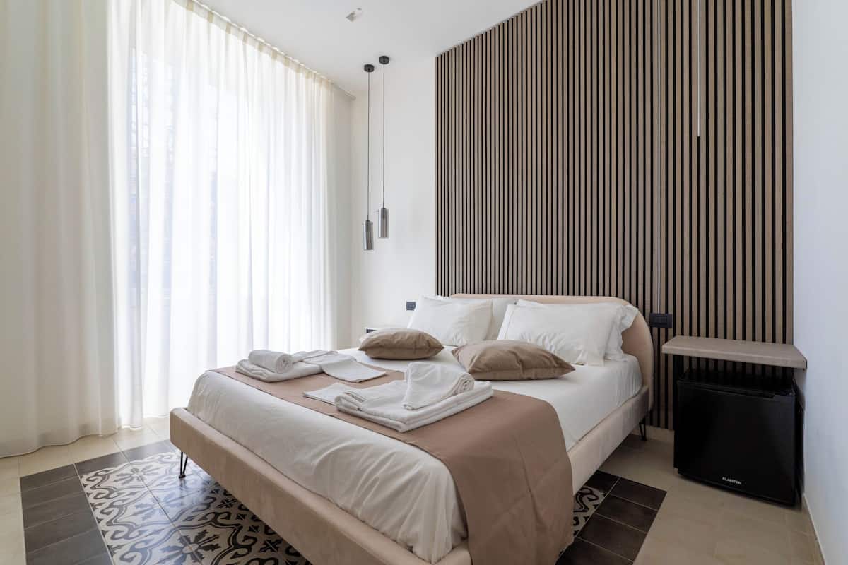 A modern bedroom with a large bed, a bedside table, a small black speaker, and a patterned rug.