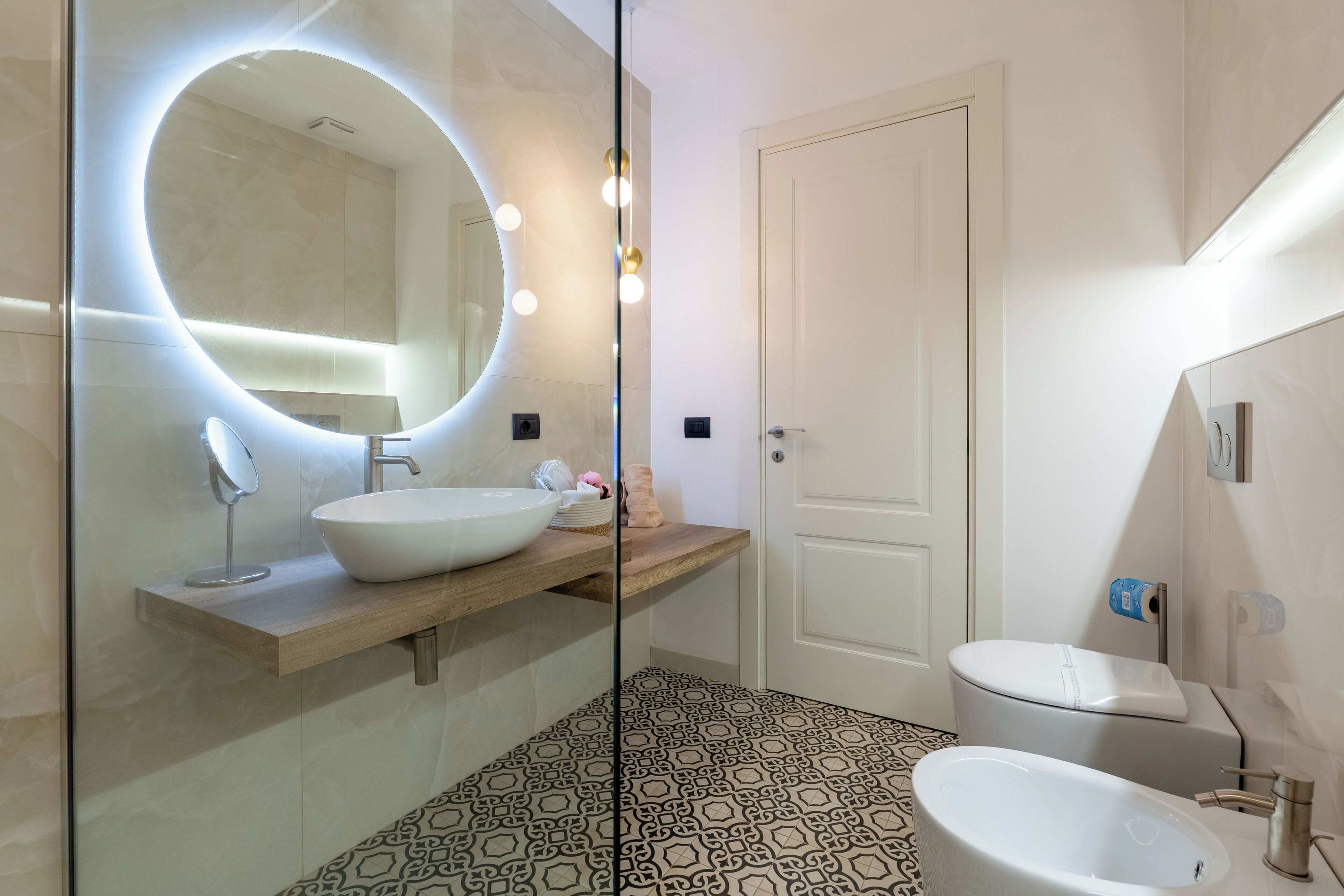 A modern bathroom with a round mirror, a wooden vanity, a freestanding bathtub, and patterned floor tiles.