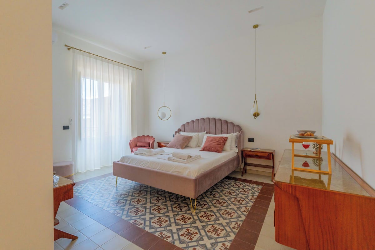 A bedroom with a bed, a bedside table, a nightstand, a mirror, and a window with curtains.