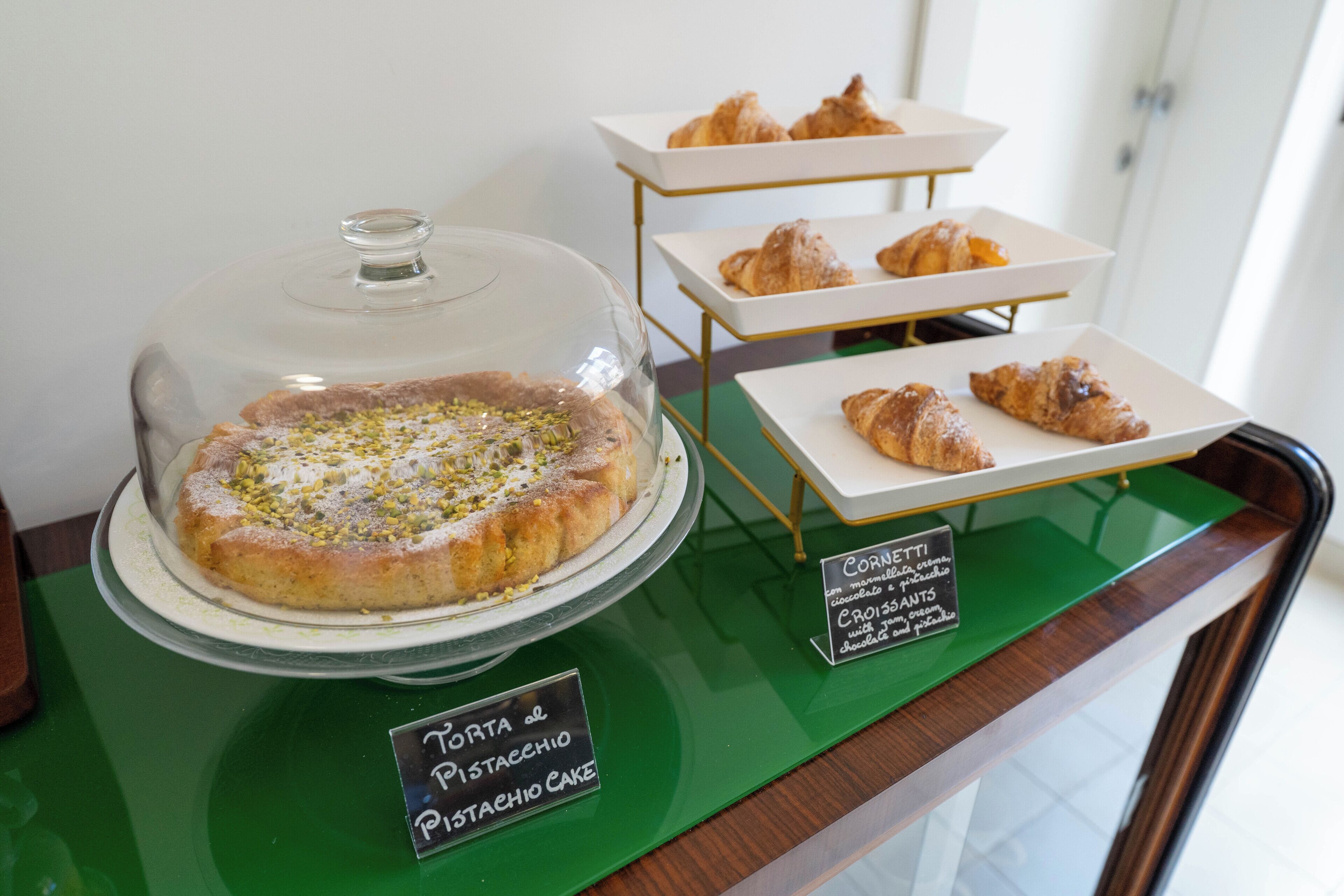 Una exhibición de pasteles y tartas, incluyendo una tarta de pistacho y croissants, sobre un mostrador verde con etiquetas.