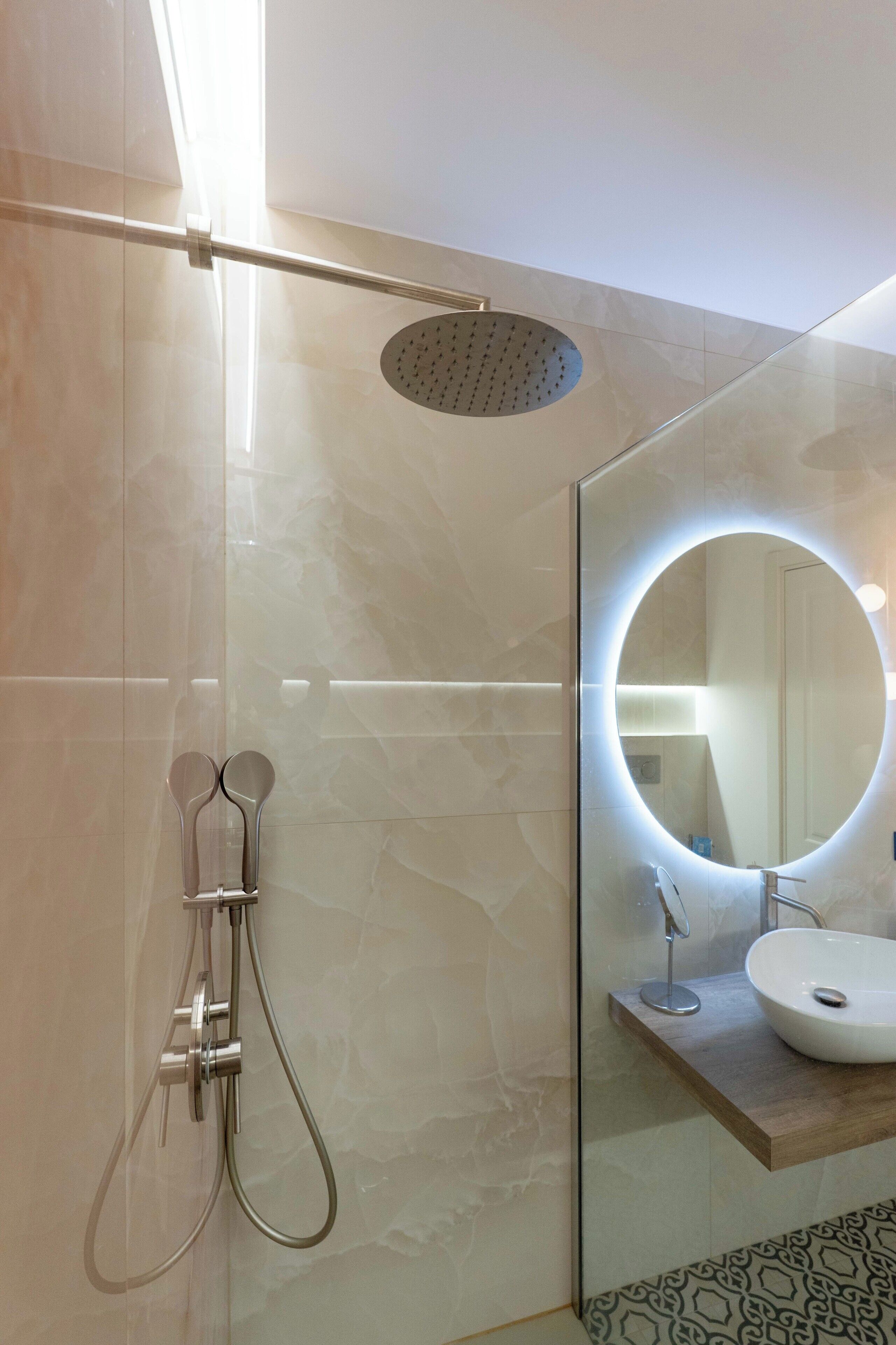 Un baño moderno con ducha envuelta en vidrio, un espejo redondo con iluminación indirecta y una encimera de madera con lavamanos.