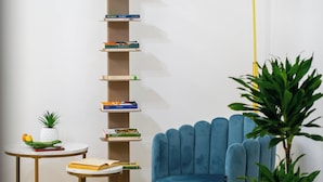 Een moderne woonkamer met een blauwe fauteuil, een hoge boekenkast, een klein rond tafeltje met een ananas en een boek, en een potplant.