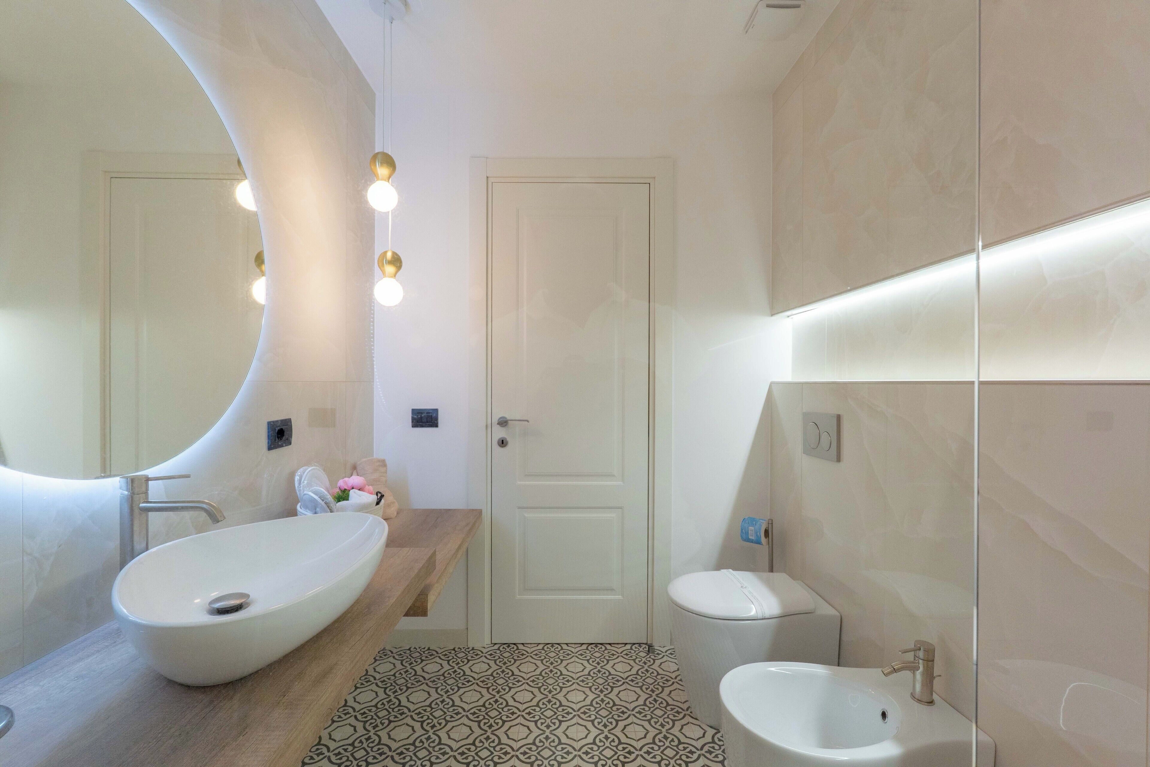 Un baño moderno con un lavamanos ovalado grande, un inodoro pequeño y un espejo con luces.