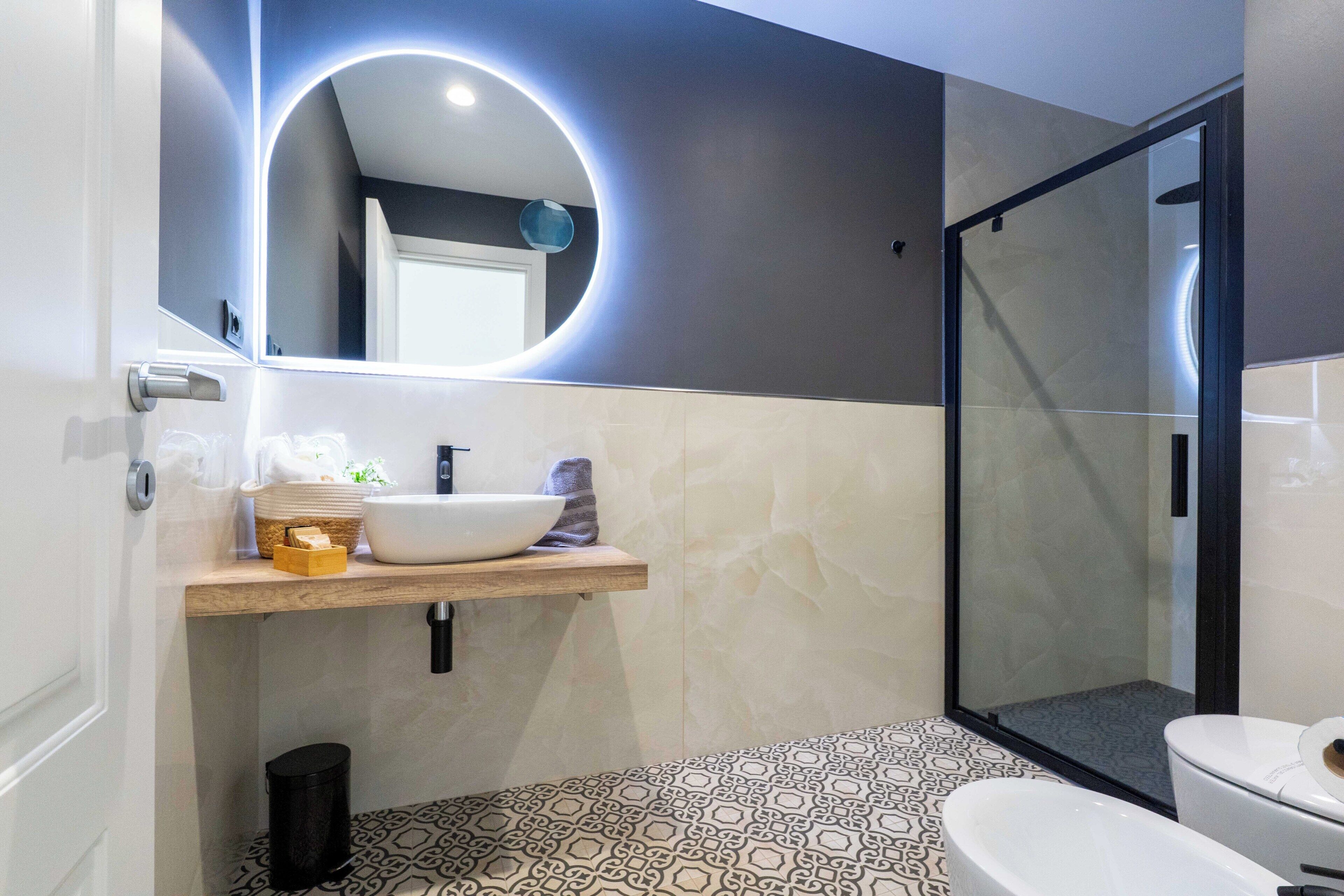 Un baño moderno con un espejo redondo, un lavabo blanco, una cabina de ducha con paredes de vidrio y baldosas de suelo estampadas.