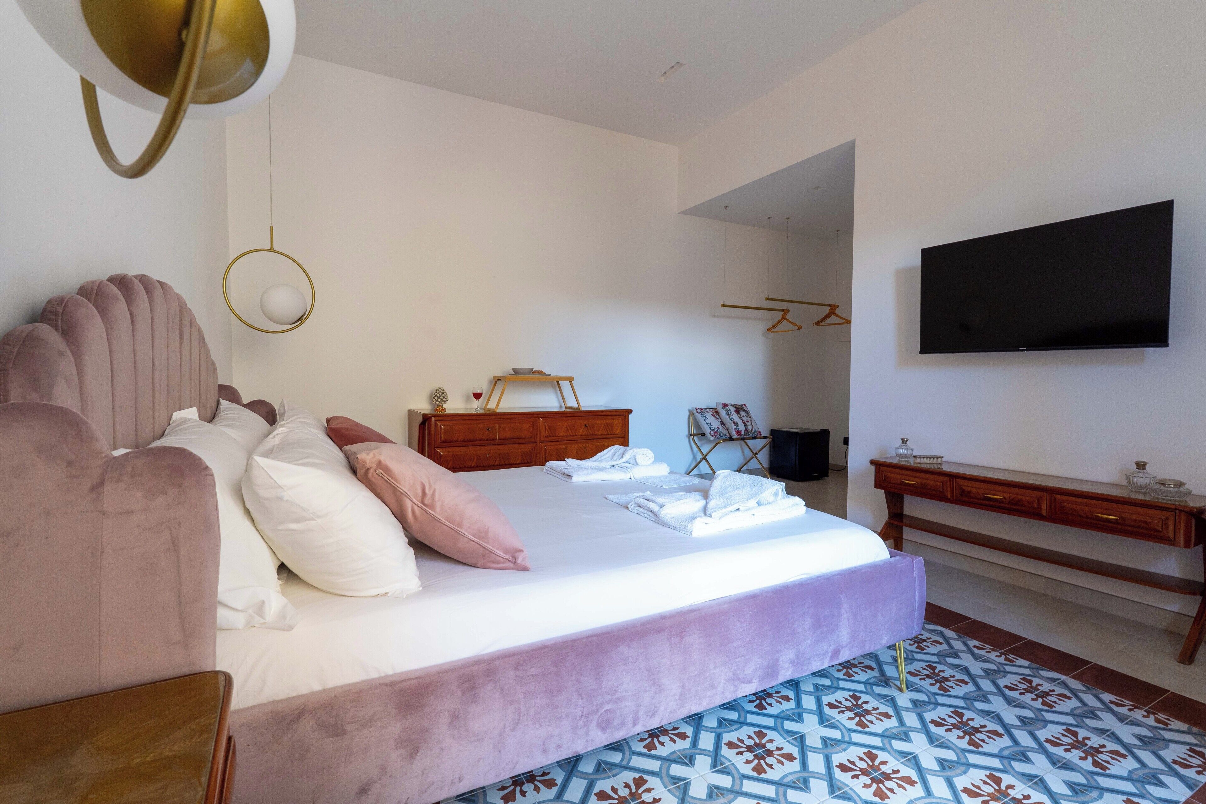 A bedroom with a large bed, a wooden dresser, a TV mounted on the wall, and a patterned rug.