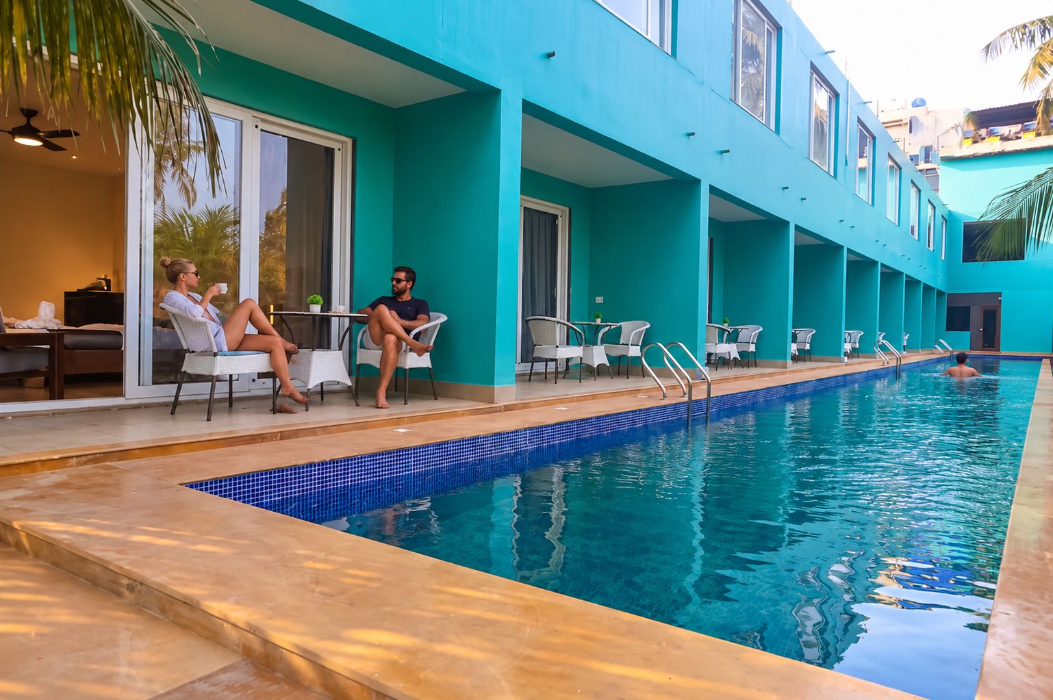 A swimming pool with lounge chairs and tables, surrounded by a building with teal walls and large windows.
