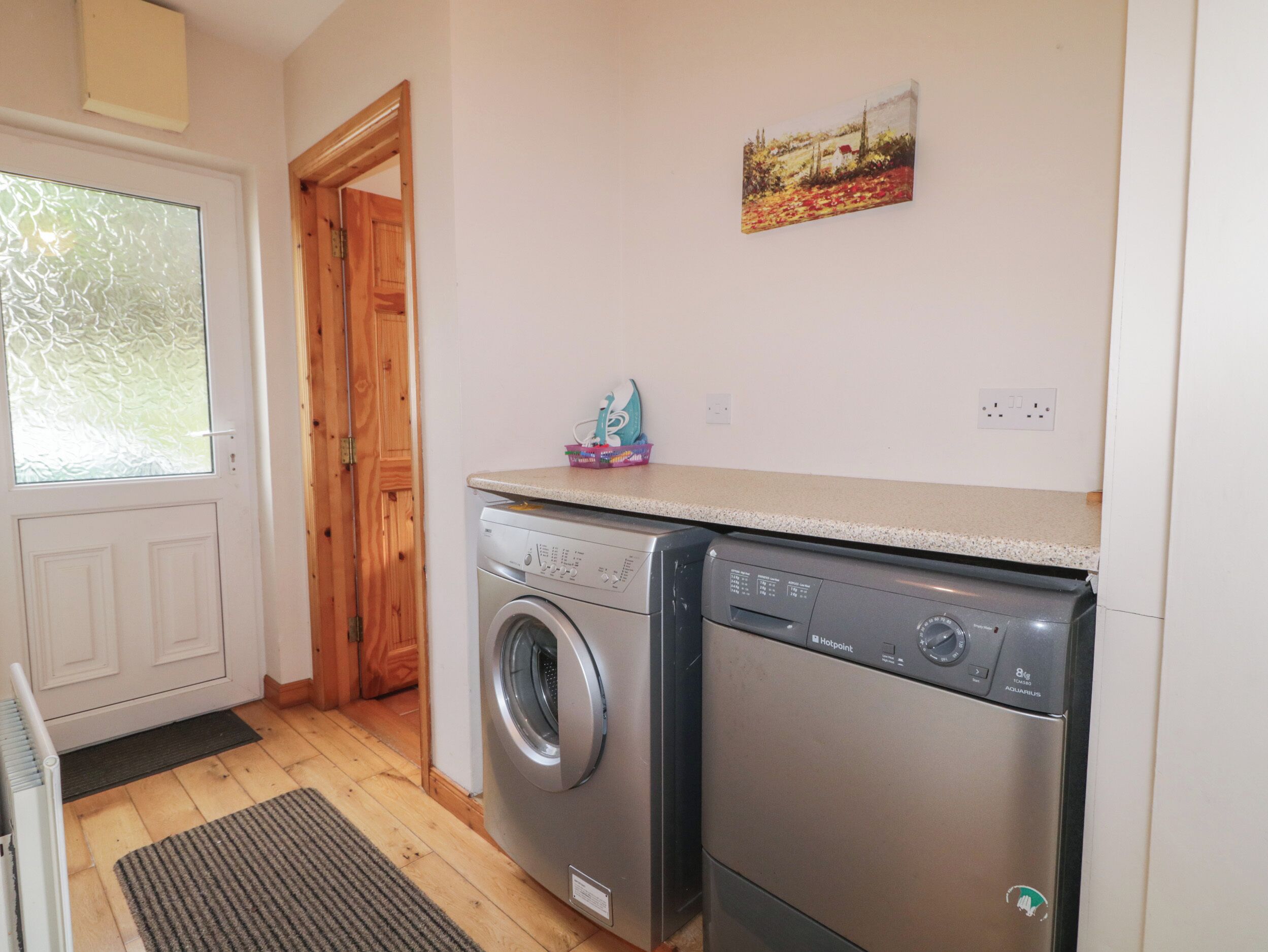 A laundry room with two washing machines, a wooden door, a window, and a painting on the wall.