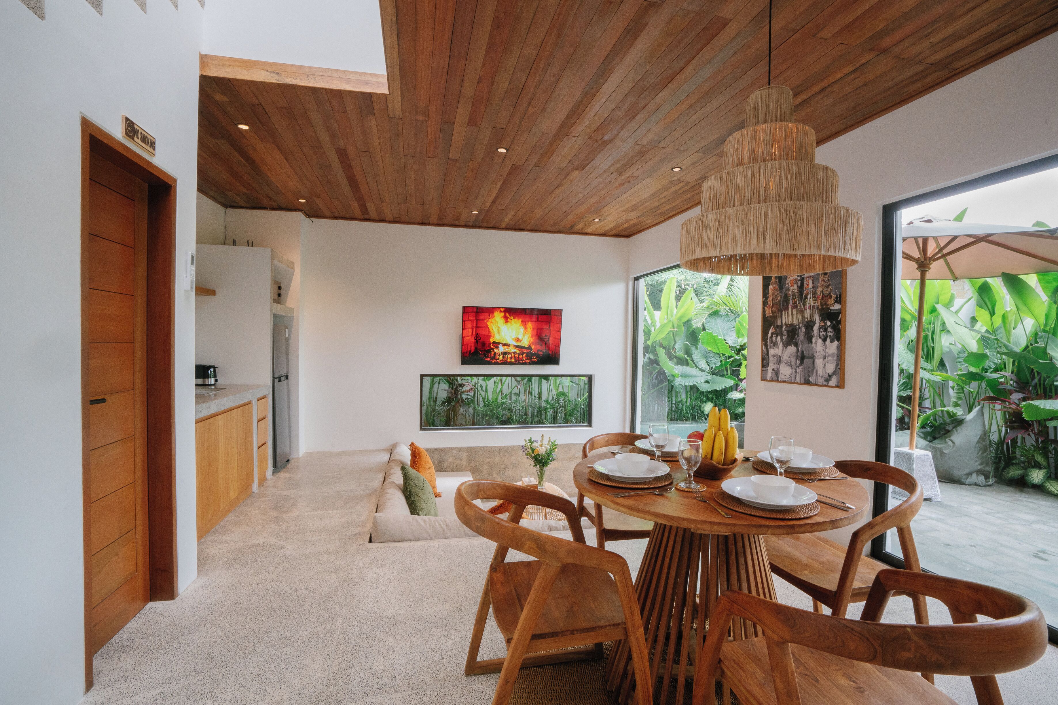 A modern dining area with a wooden table and chairs, a large window with a view of greenery, and a ceiling with wooden beams.