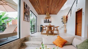 A modern living room with a wooden ceiling, a dining area with a round table and chairs, a sofa with cushions, and a staircase leading to another level. - The Reef Villas (Pecatu)