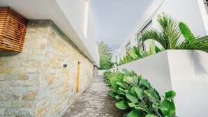 A narrow, paved alleyway flanked by white buildings with stone walls on one side and a white wall with green plants on the other. - The Reef Villas (Pecatu)