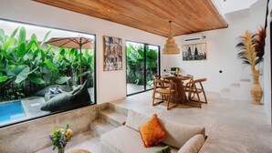 A modern living room with a large window, a dining area, and a staircase. - The Reef Villas (Pecatu)