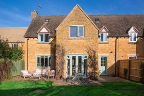 Une maison en pierre à deux étages, dotée d’une entrée centrale, d’un patio couvert avec des meubles d’extérieur et d’une cour arrière clôturée.