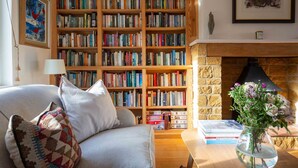A cozy living room with a large wooden bookshelf filled with books, a comfortable sofa with cushions, a small table with a vase of flowers, and a fireplace. - fabulous and homely dog friendly holiday cottage  - Kingsley House (Draycott)
