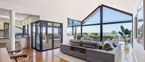 A modern living room with a large glass wall, a grey sofa, wooden flooring, and a view of the ocean.