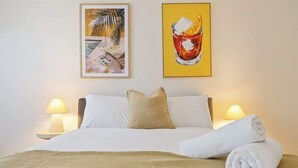 A hotel room with a bed, two bedside lamps, a rolled towel, and two framed artworks on the wall.