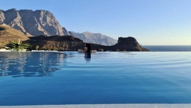A clear blue infinity pool with a person standing in the water, surrounded by rocky mountains and a clear sky. - Vivelorural Agaete Serenity garden in El Turmán (Agaete)
