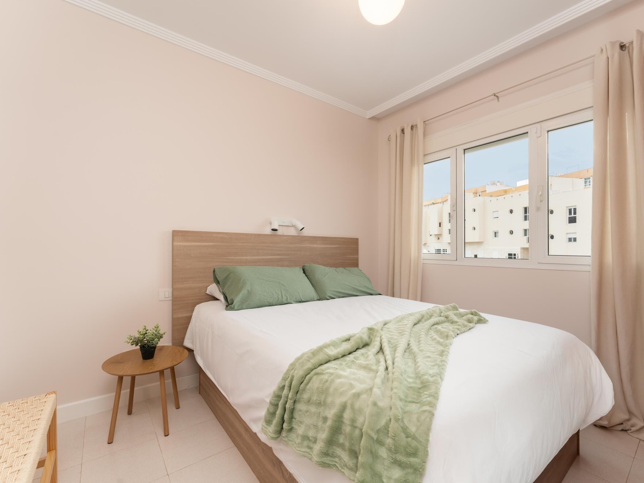 A neatly arranged bedroom with a bed, a small wooden table with a plant, and a view of buildings through the window.