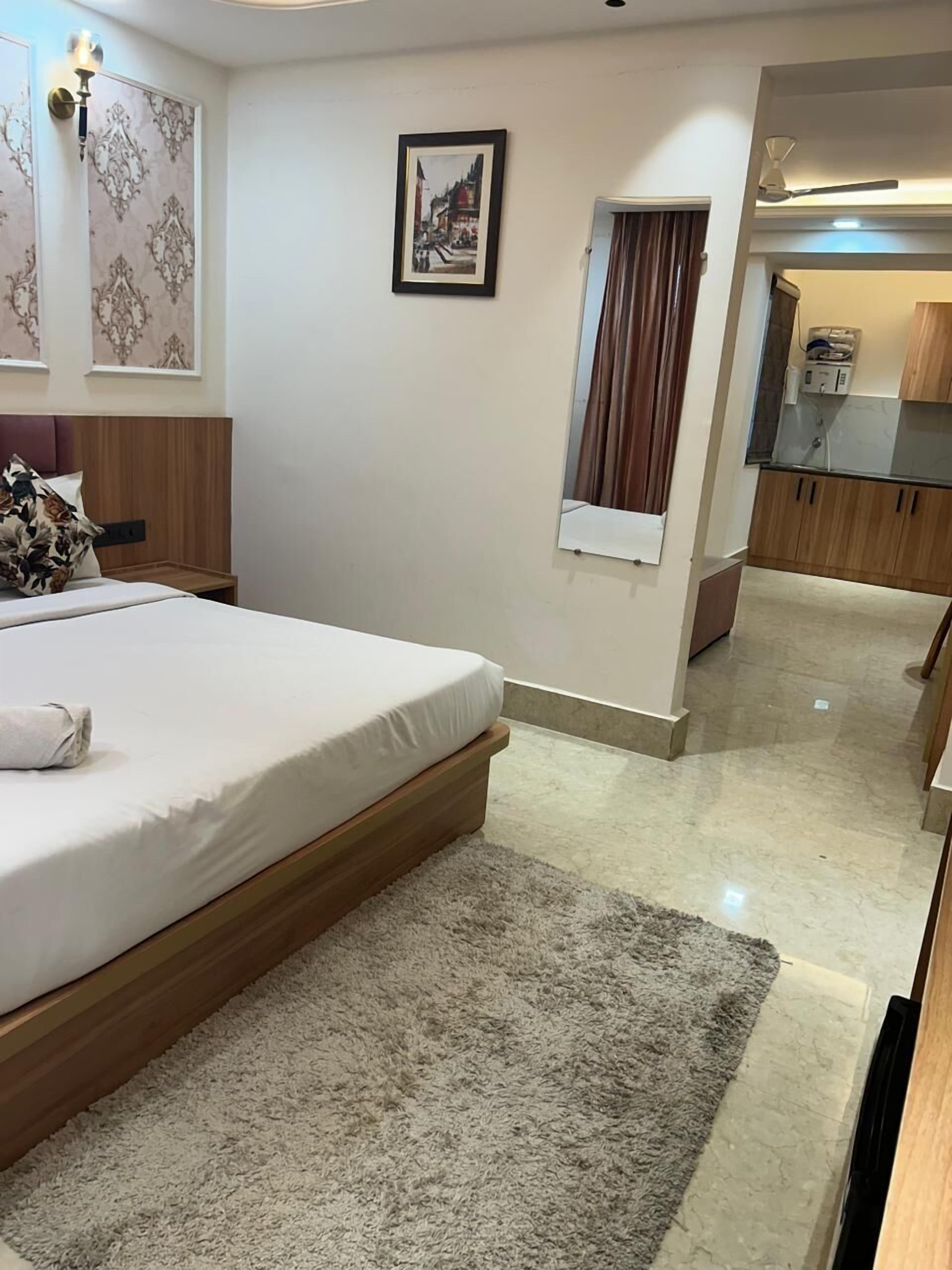 A hotel room with a bed, a small window, a mirror, a wall-mounted light fixture, and a view of the kitchen.