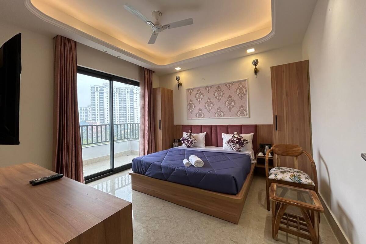 A modern hotel room with a large bed, a desk, a chair, and a balcony with a view of high-rise buildings.