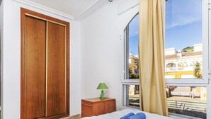 A bedroom with a large window, a bed with white bedding, a green lamp on a wooden nightstand, and a wooden wardrobe.