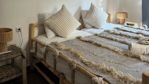 Une chambre avec un lit en bois, du linge de lit blanc, une décoration murale tissée et un miroir rond.