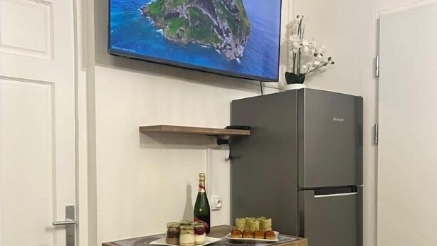 A compact kitchen with a wooden table, two black chairs, a refrigerator, and a television mounted on the wall displaying a scenic view. - Lovely studio, ideal for a couple!  (Schoelcher)